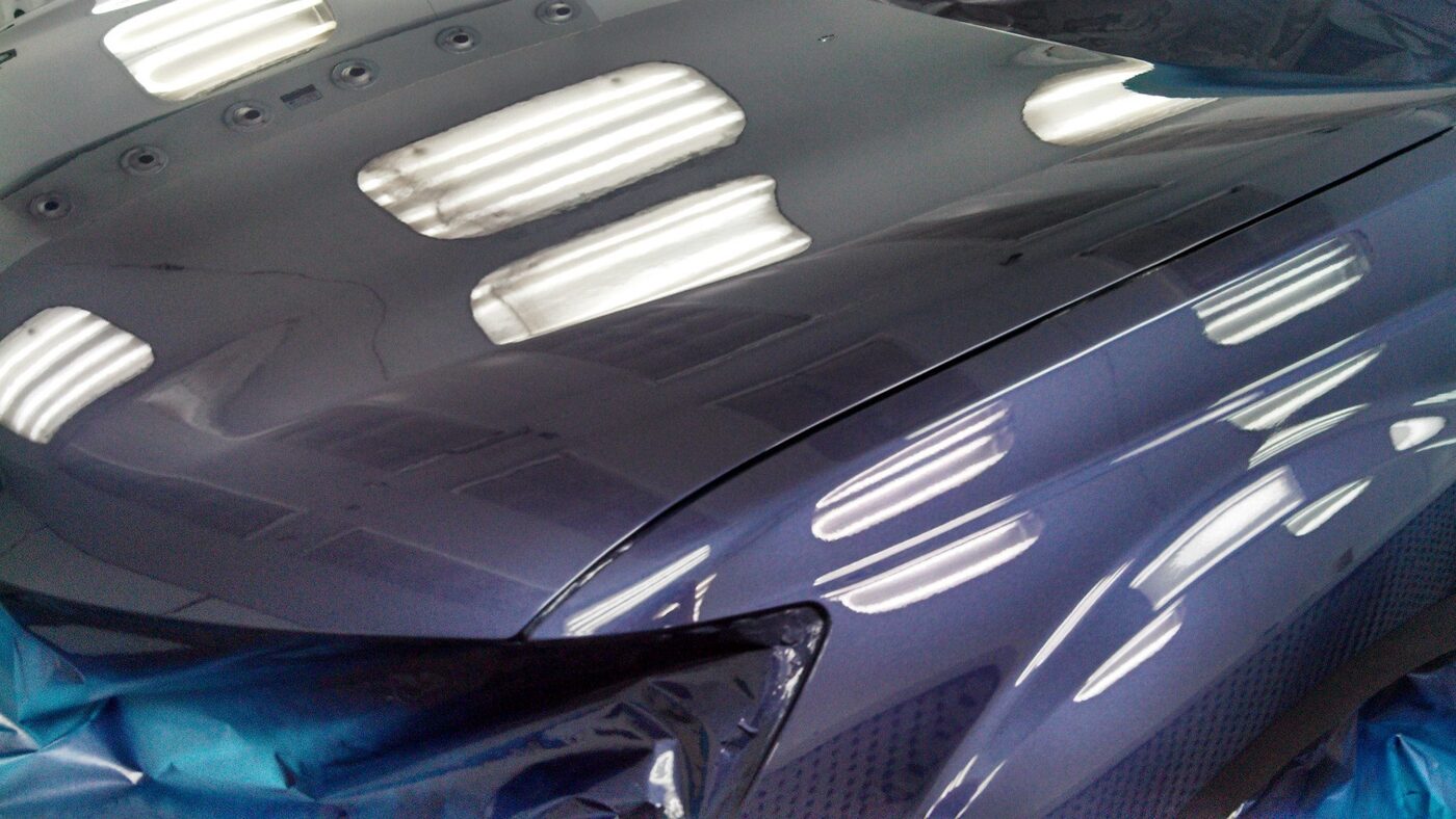 A freshly refinished car panel inside the Malibu Collision paint booth, the booth lights reflected down the length of the hood